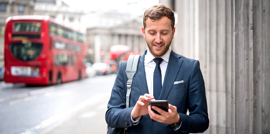 Yurt dışında arka planda kırmızı çift katlı bir otobüs geçerken akıllı telefonuna bakıp gülümseyen takım elbiseli adam