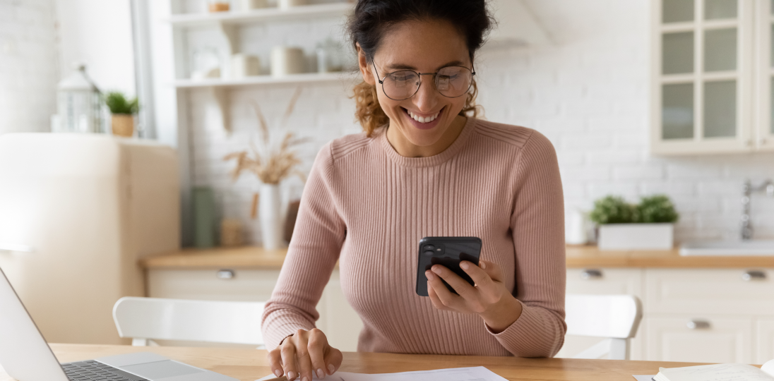 A smiling young woman sitting at a table with a phone in one hand and a calculator in the other